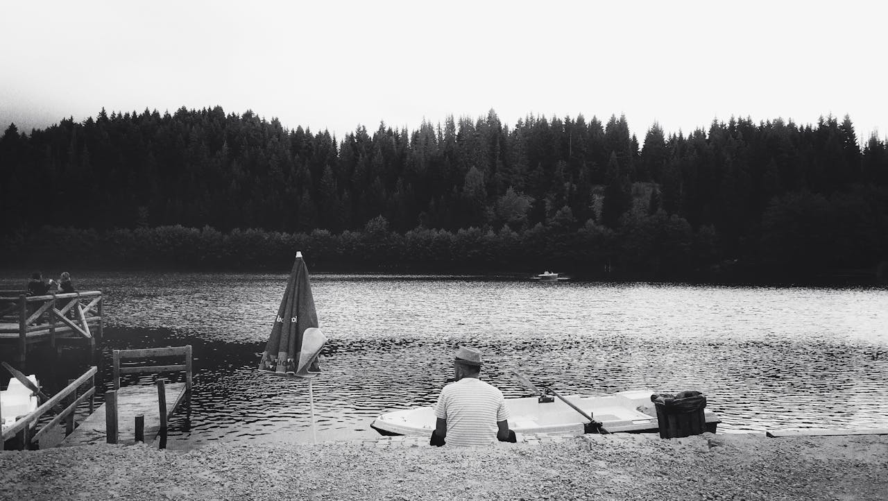 Peaceful lakeside view with a lone figure, boat, and dense forest in black and white.