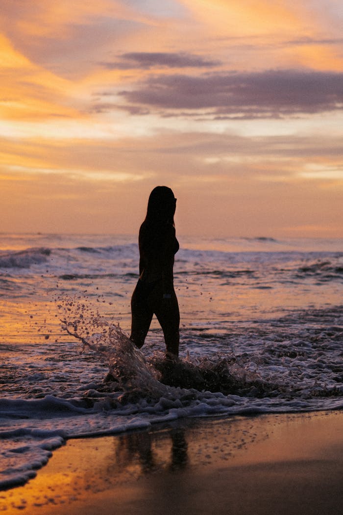 Silhouette of a woman standing in waves at sunset, peaceful beach scene.